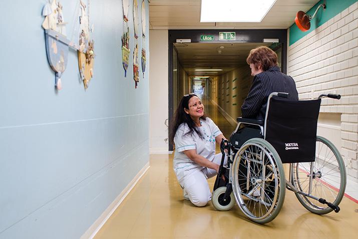 Potilas istuu pyörätuolissa, potilaskuljettaja asettelee potilaan jalkoja puörätuolin kannattimille