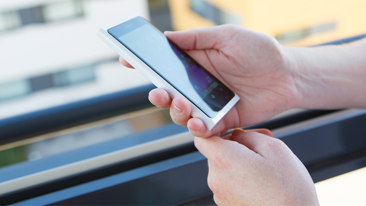 Phone in the hand of an adult, in the background a large window and a block of flats in summer sun.