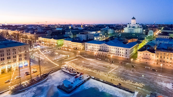 Ilmakuva talvisen Helsingin ydinkeskustasta iltahämärän aikaan, sininen taivas, keltaiset valot, taustalla tuomiokirkko.