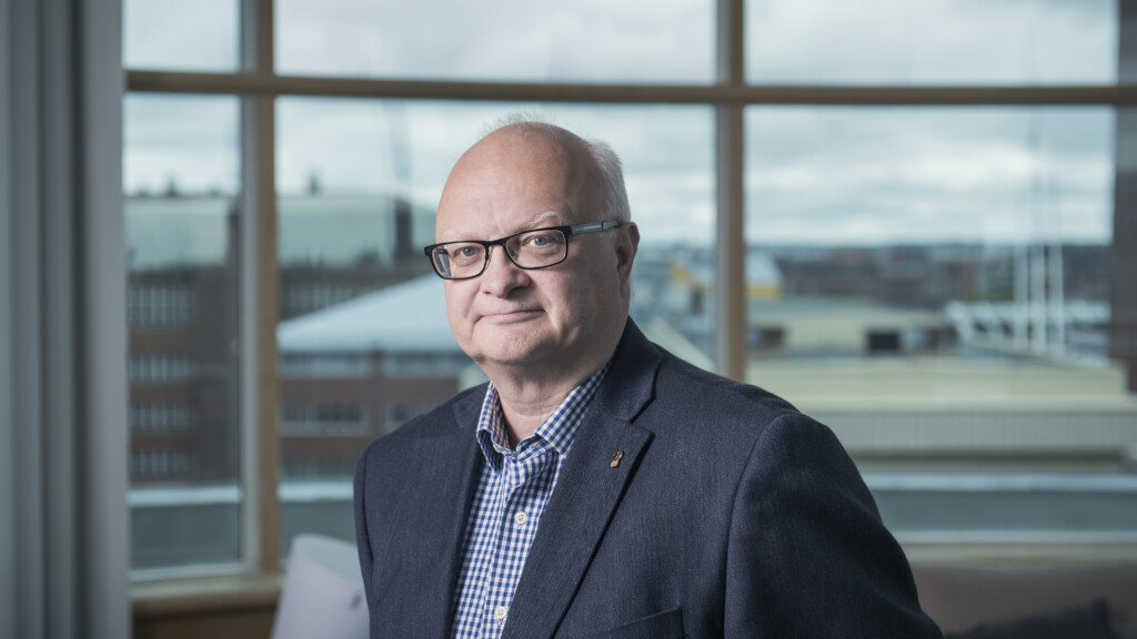 Mies seisoo neuvotteluhuoneessa tumma puku päällä, silmälasit päässä.