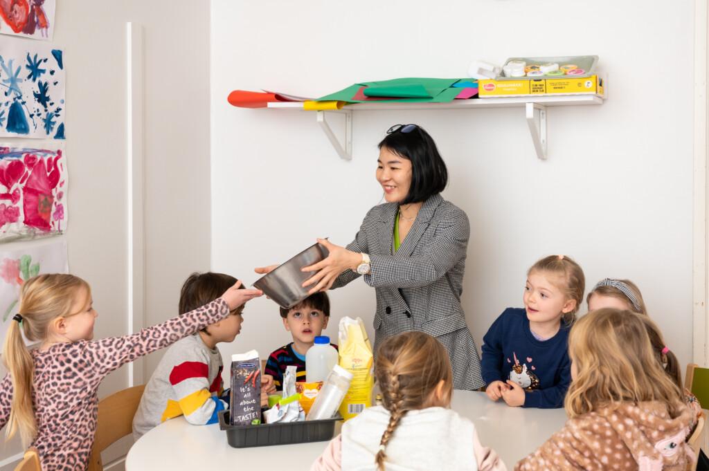 En vuxen och åtta barn runt ett bord. Den vuxna räcker ett av barnen en skål. På väggen syns teckningar och en hylla.