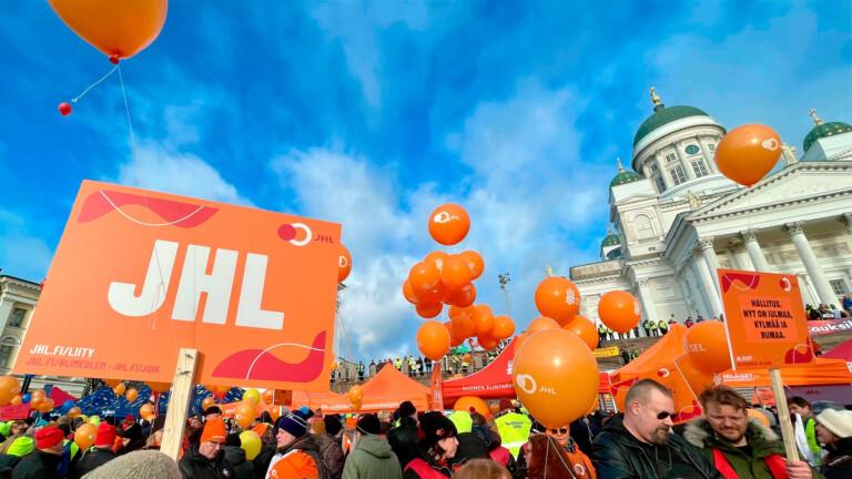 Orange strike sign with JHL written in white, sunny Senate Square in the background, white clouds in the sky, Helsinki Cathedral in the background.