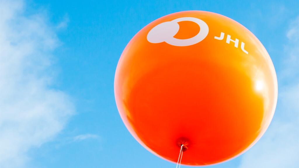An orange balloon with a white JHL print, floating against a backdrop of bright blue sky and white clouds.