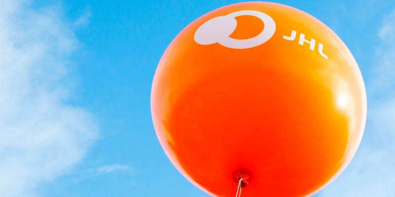 Oranssi ilmapallo, jossa valkoisella teksti JHL, taustalla poutapilvinen taivas.