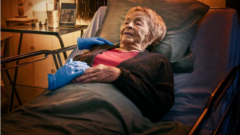 Vanha ihminen makaa sairaalasängyssä. Hoitajan kädet koskettavat henkilöä pehmeästi. Hoitajasta näkyy vain työhanskat, muuten hän on näkymätön.