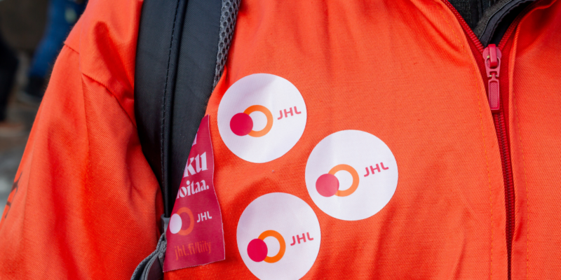 Ammattiliitto JHL:n nuorella jäsenellä on päällä oranssit opiskelijahaalarit. Lähikuva haalarin etuosasta. Runnukseen on liimattu kolme JHL:n tarraa ja yksi Kyllä joku hoitaa -tarra.
