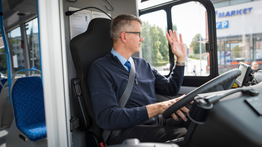 En busschaufför sitter vid ratten, tittar ut igenom fönstret och lyfter sin hand i en hälsning.