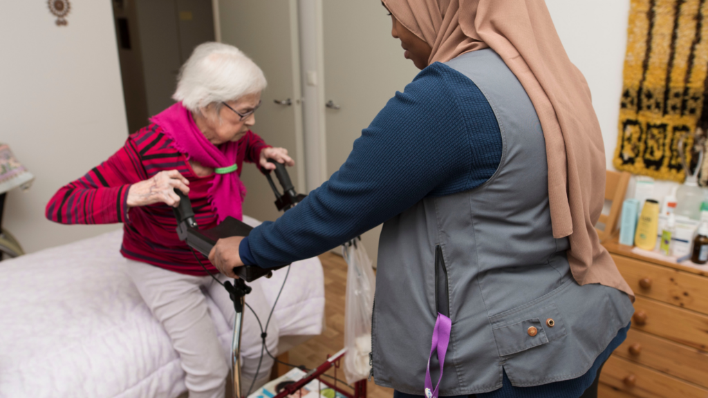 En äldre person håller på att sätta sig på en säng och stöder sina händer på en rollators handtag. En annan person håller rollatorn stadigt på plats.