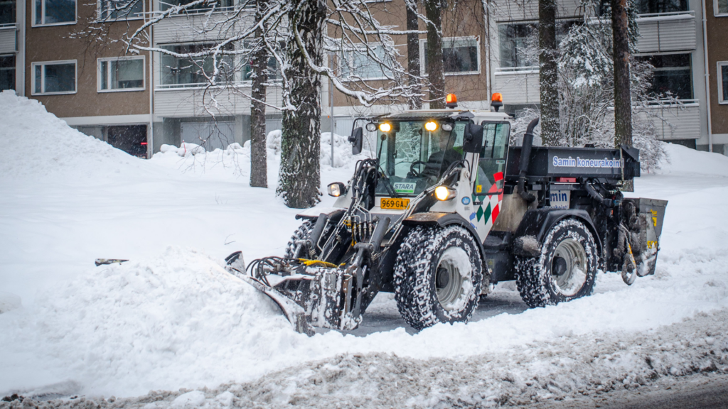 En plog plogar en trottoar i ett snöigt stadslandskap.