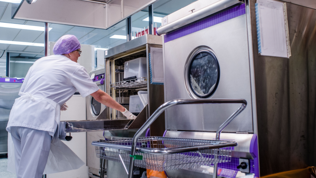 En person i vita laboratoriekläder placerar lådor av plast i en maskin för rengöring.